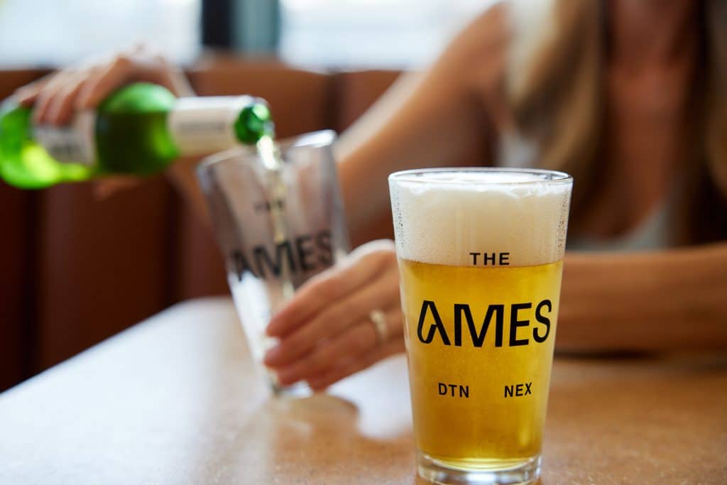 Lifestyle photo of a woman pouring beer into a branded 'The Ames' glass cup